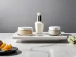 Tres envases de productos de belleza, dos tarros y una botella con dosificador, sobre una bandeja de mármol blanco. Fondo gris y encimera de mármol veteado con naranjas y flores.